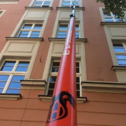 Czerwony wysięgnik z logo firmy sięga do okien kamienicy o jasnej fasadzie, widoczne odbicia nieba w szybach.