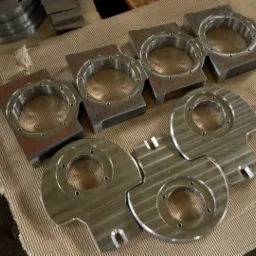 Grupowe ujęcie ośmiu obrobionych metalowych elementów z centralnym otworem i otworami montażowymi, widoczne ślady obróbki skrawaniem, ułożone na kartonowej powierzchni.