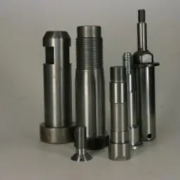 Zestaw precyzyjnie obrobionych, metalowych elementów o cylindrycznym kształcie i zróżnicowanych rozmiarach, ustawionych na jasnym tle. Widoczne gwinty, otwory i schodkowe przekroje.