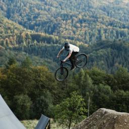 PN Patryk Nawrocki - Mężczyzna w kasku wykonuje skok na rowerze górskim na tle zalesionych wzgórz i rampy.