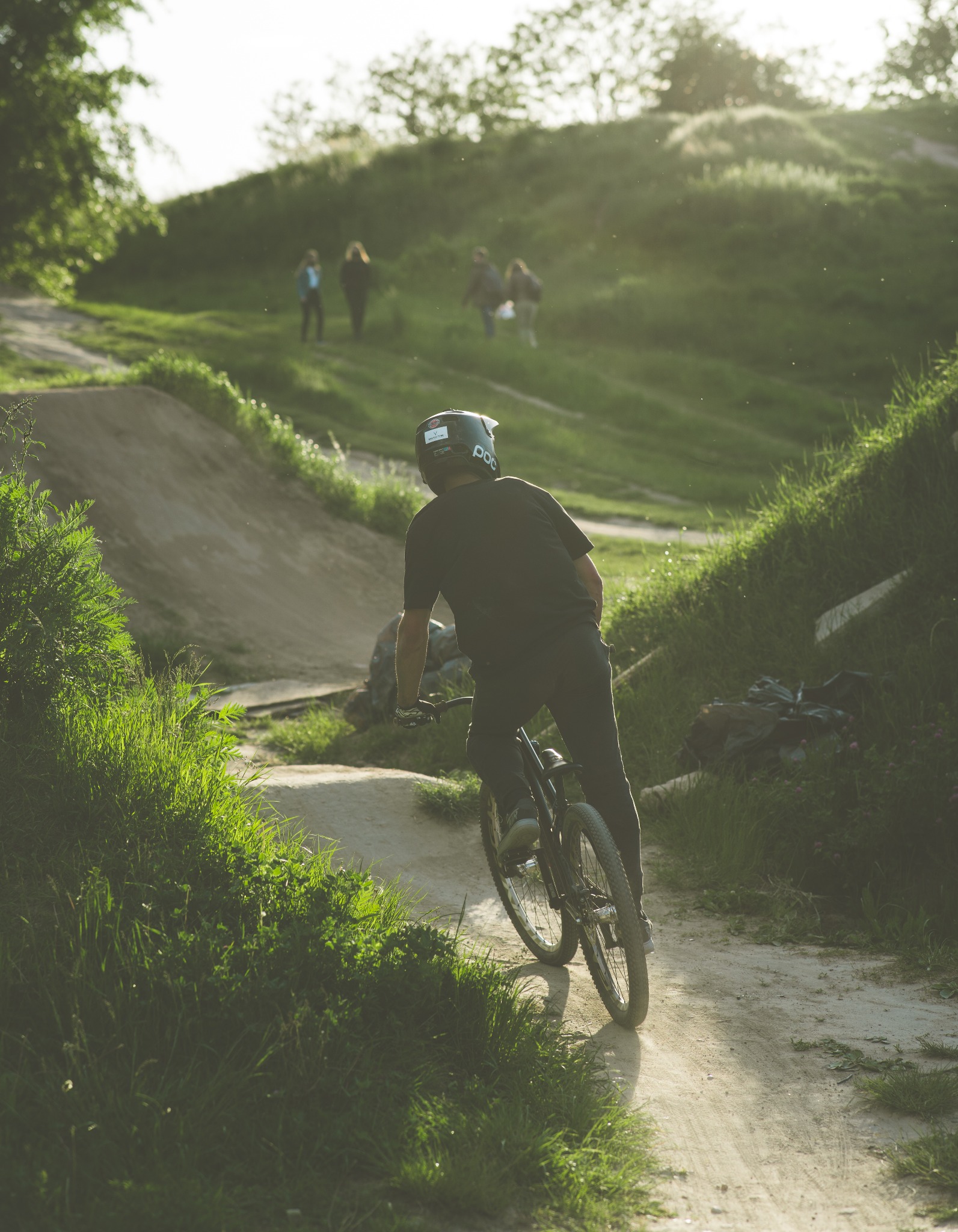 Mężczyzna w kasku i czarnym stroju jedzie rowerem górskim po ziemnym torze z przeszkodami, w tle zielone wzgórze i grupa osób.