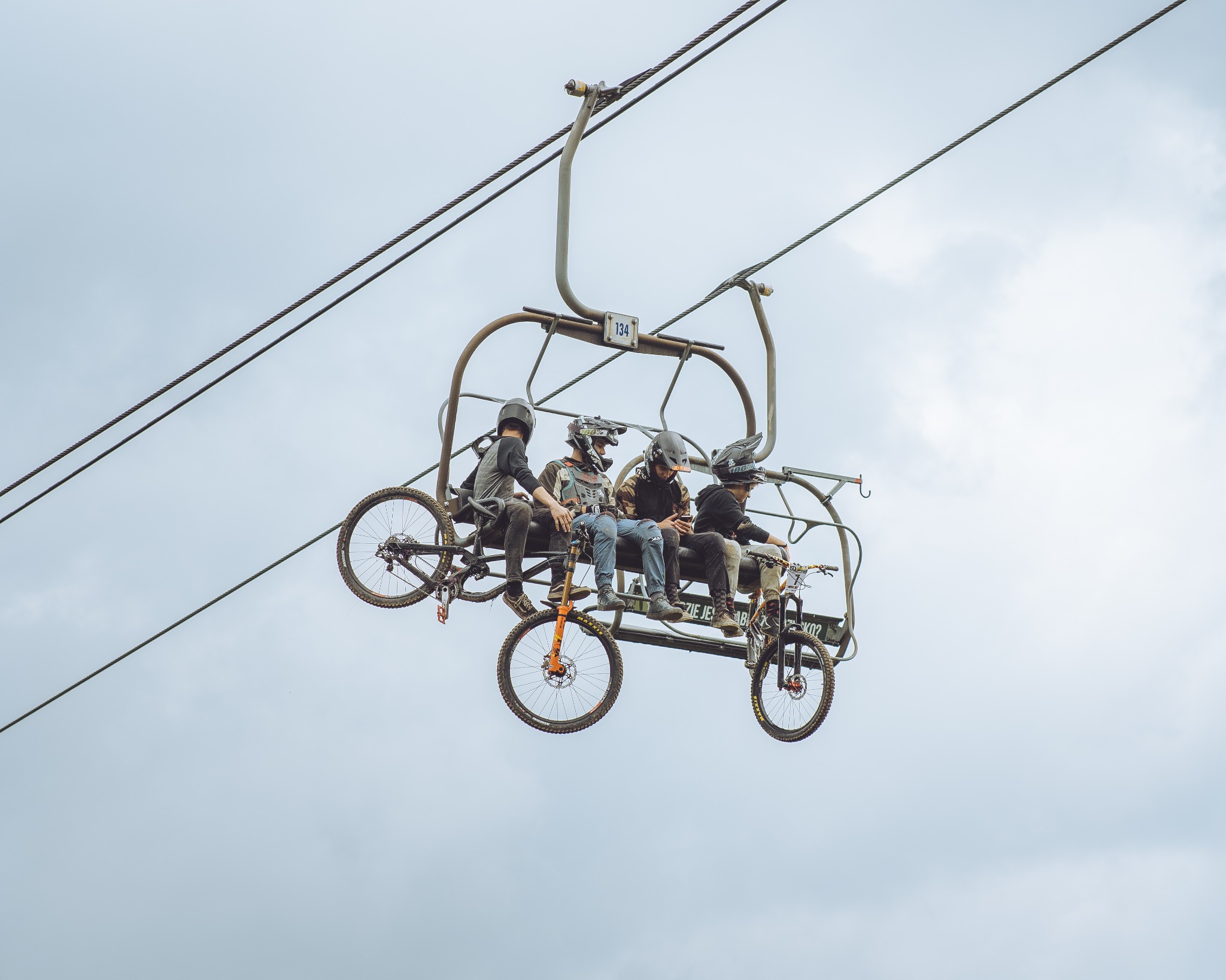 Czterech rowerzystów w kaskach i ochraniaczach siedzi na wyciągu krzesełkowym z rowerami górskimi, widok z dołu na tle pochmurnego nieba.