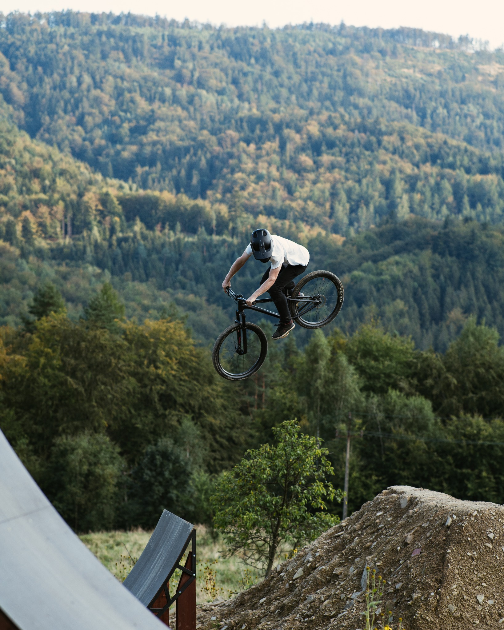 Mężczyzna w kasku wykonuje skok na rowerze górskim na tle zalesionych wzgórz i rampy.