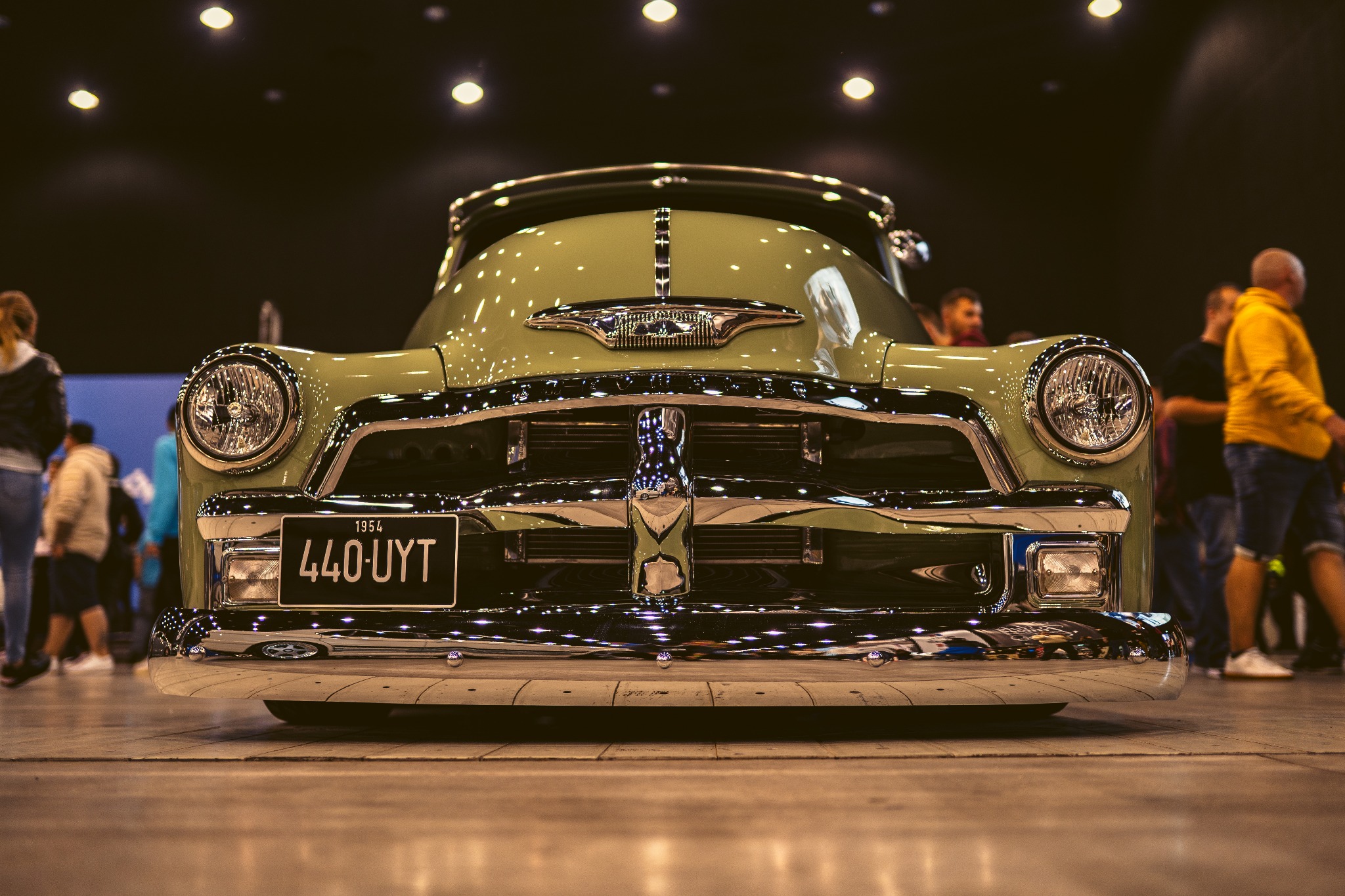 Przód odrestaurowanego, jasnozielonego samochodu z rocznika 1954 z chromowanymi elementami, widoczna tablica rejestracyjna 440-UYT, na tle rozmytych postaci i oświetlenia hali.