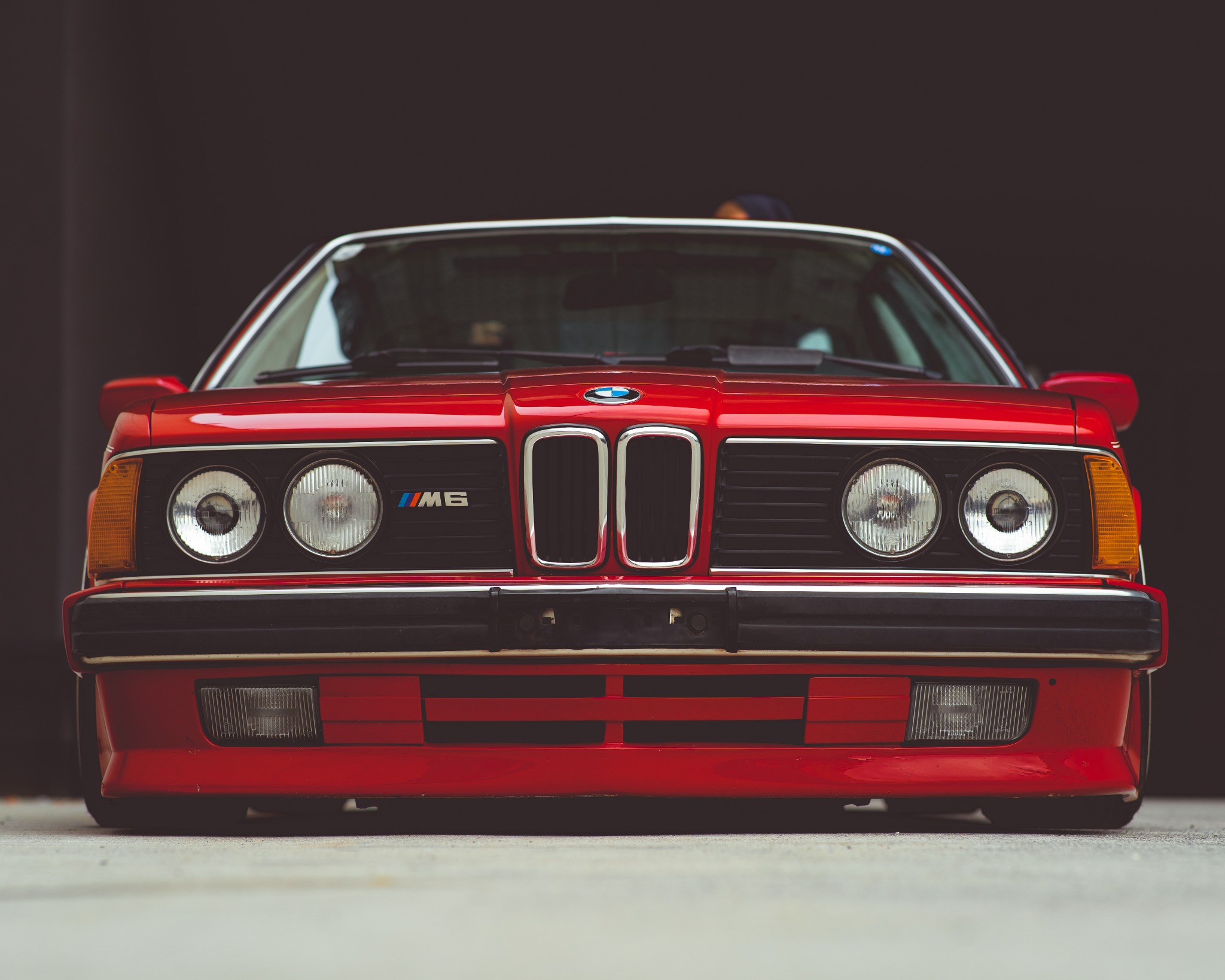 Czerwone BMW M6 E24, widok z przodu, charakterystyczna atrapa chłodnicy z chromowanymi nerkami, okrągłe reflektory, emblemat M6 na grillu, niski profil, ciemne tło.