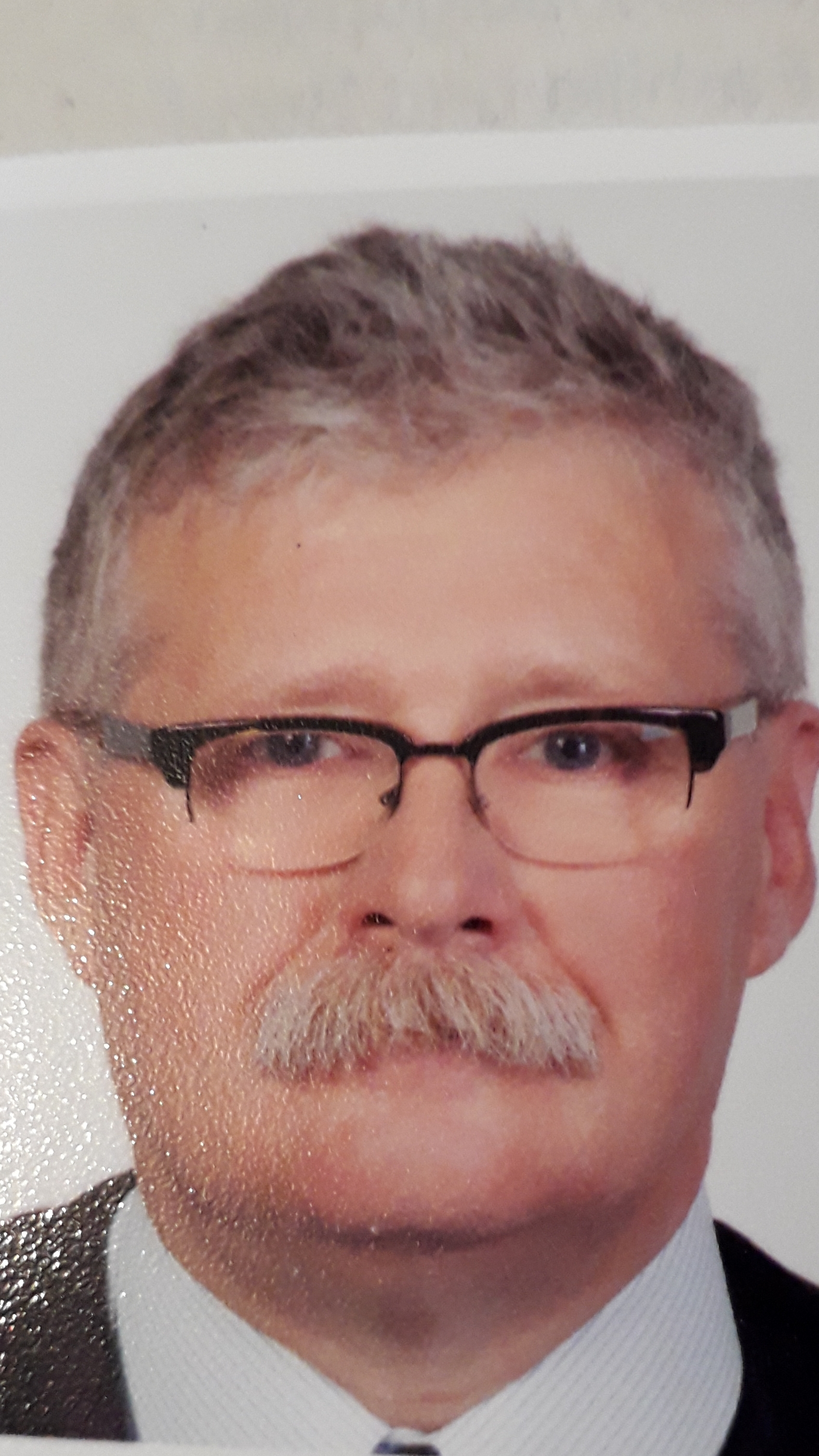 Portret starszego mężczyzny w okularach i koszuli z krawatem, z widocznymi zmarszczkami i szpakowatymi włosami, na jasnym tle.