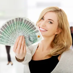 Uśmiechnięta blondynka trzyma wachlarz z banknotów 100 zł na rozmytym tle biurowym z dwoma mężczyznami.