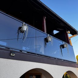 Alu Prospect - Szklana balustrada balkonowa z wiszącymi doniczkami, widok z dołu na tle błękitnego nieba, z widocznymi detalami architektonicznymi budynku w stylu klasycznym.