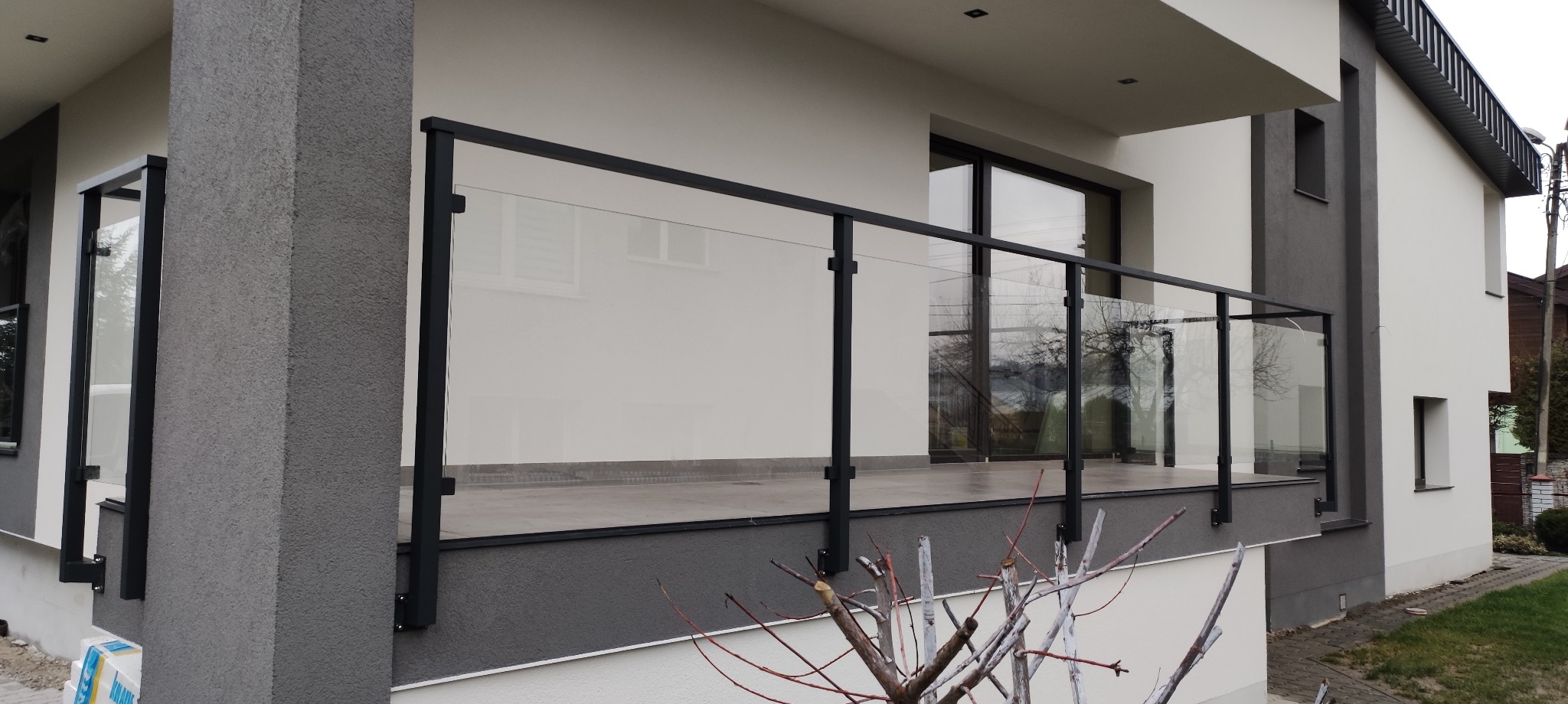 Nowoczesna balustrada szklana z czarnymi profilami na balkonie domu jednorodzinnego. Widok na elewację z oknami. Minimalistyczny design, jasna kolorystyka.