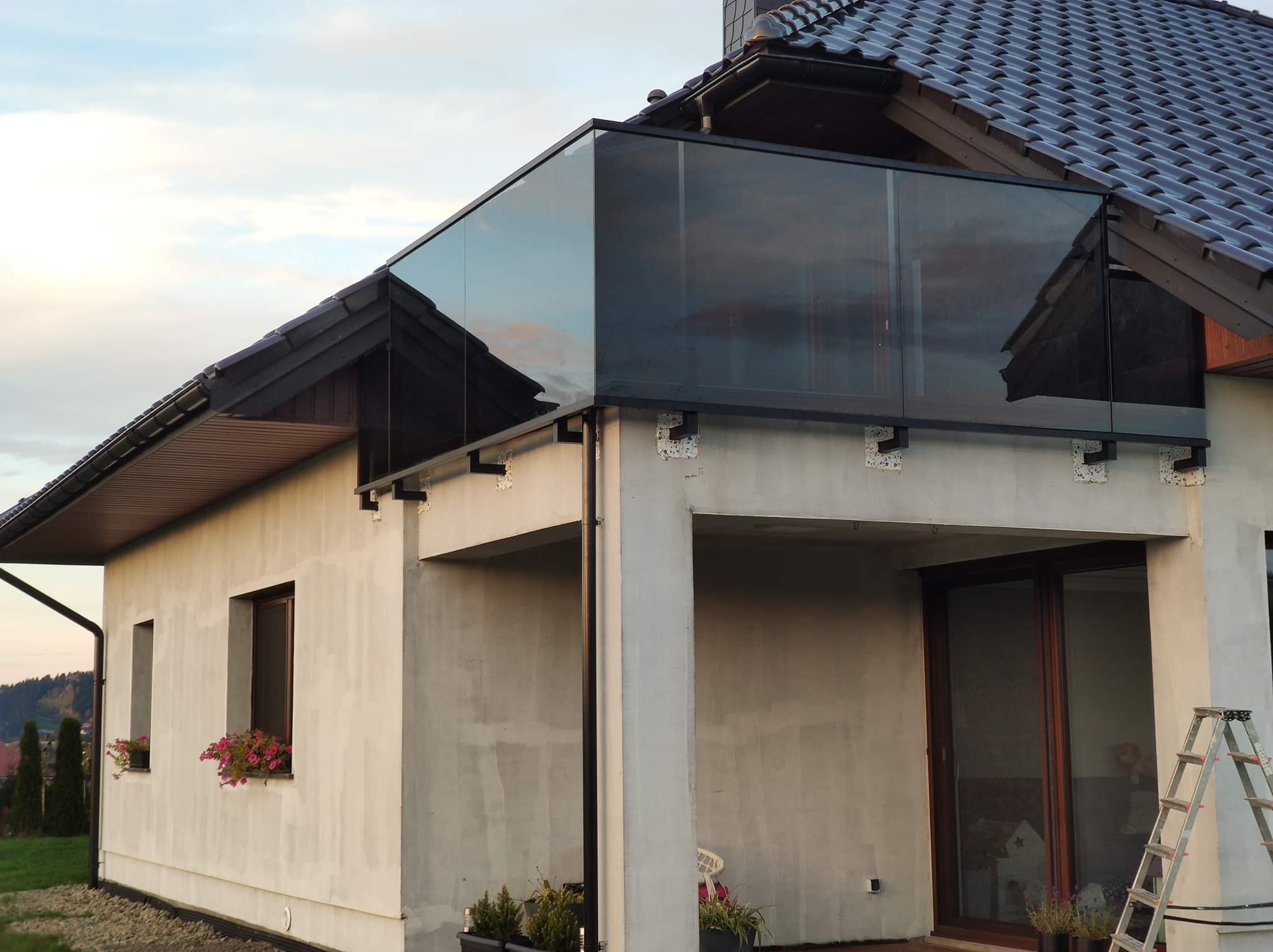 Szklana balustrada na balkonie domu jednorodzinnego, widok z dołu, z drabinką opartą o ścianę. Minimalistyczny design, nowoczesne wykończenie, jasna elewacja.