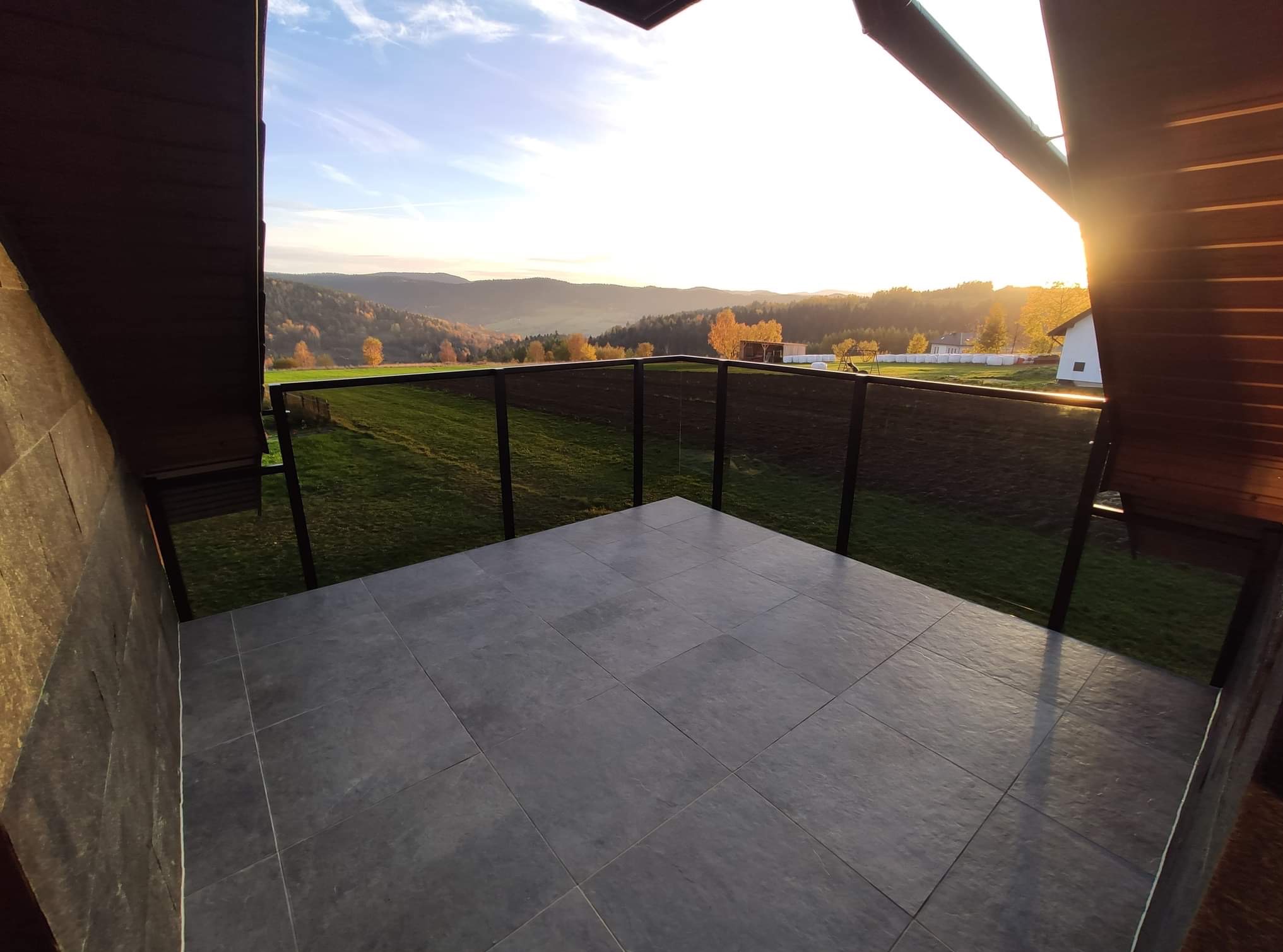 Szklana balustrada na balkonie z szarymi płytkami, widok na wzgórza w ciepłym świetle zachodzącego słońca. Nowoczesny design, otoczenie zieleni i drzew.