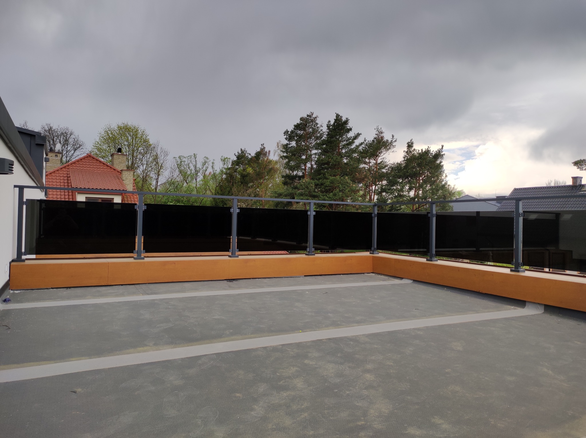 Nowoczesna balustrada szklana z grafitowymi słupkami na tarasie, z widokiem na drzewa i pochmurne niebo w tle. Minimalistyczny design i funkcjonalność.