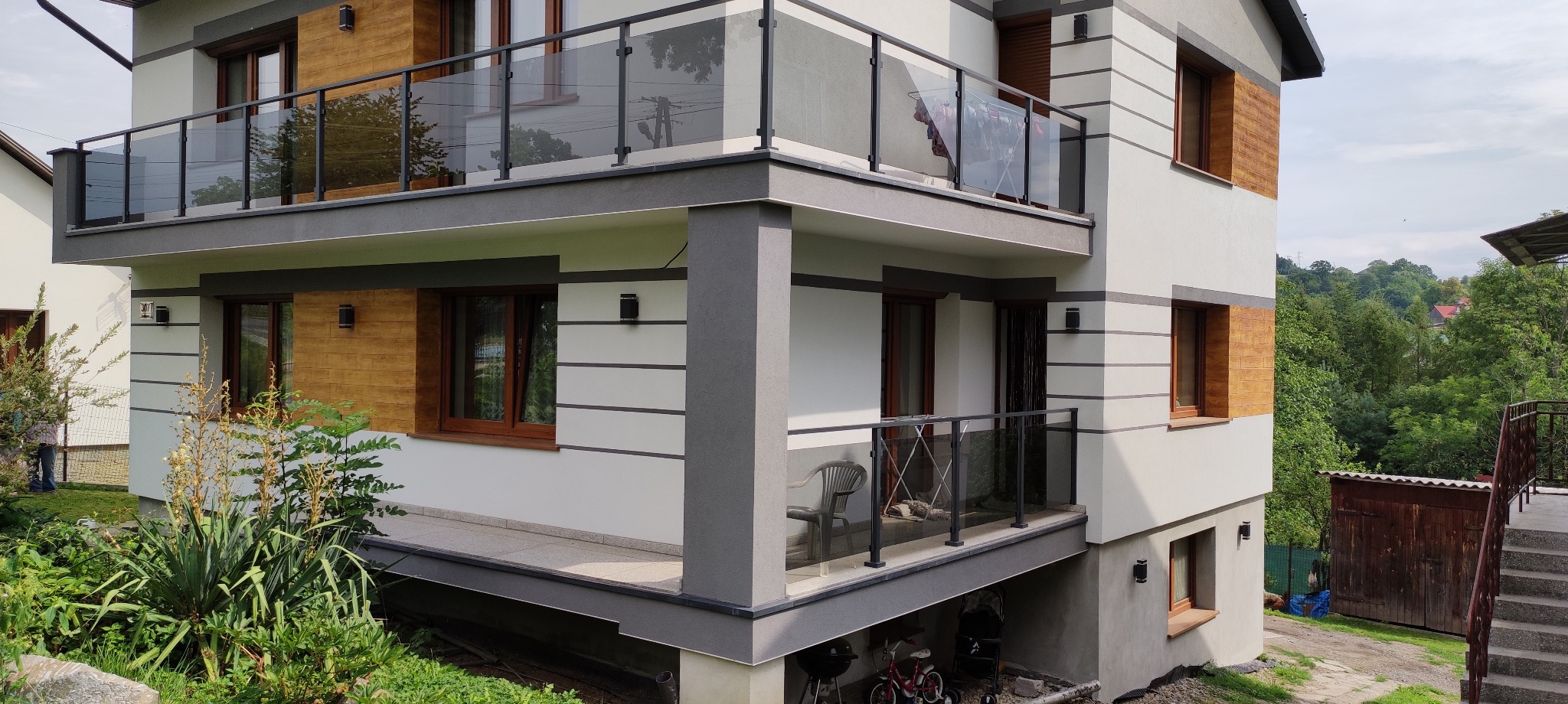 Nowoczesny dom z balkonami zabezpieczonymi szkłem hartowanym i czarnymi ramami, widok z dołu na fasadę budynku i otoczenie zieleni.