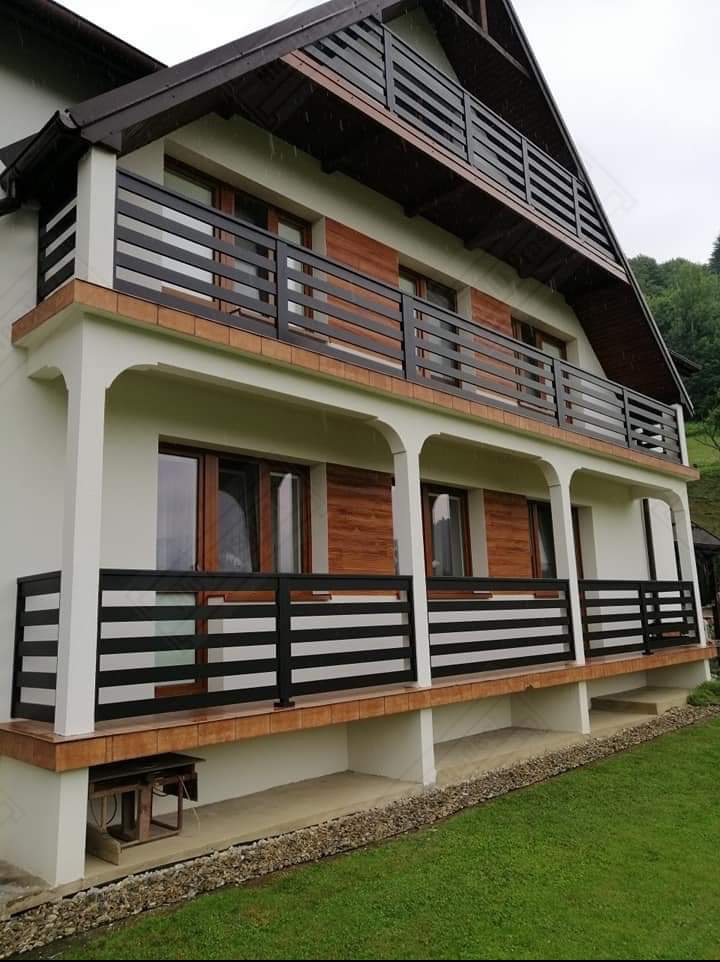 Dwupiętrowy dom z balkonami zabezpieczonymi czarnymi, poziomymi balustradami. Widoczne drewniane okiennice i tło z zielenią.