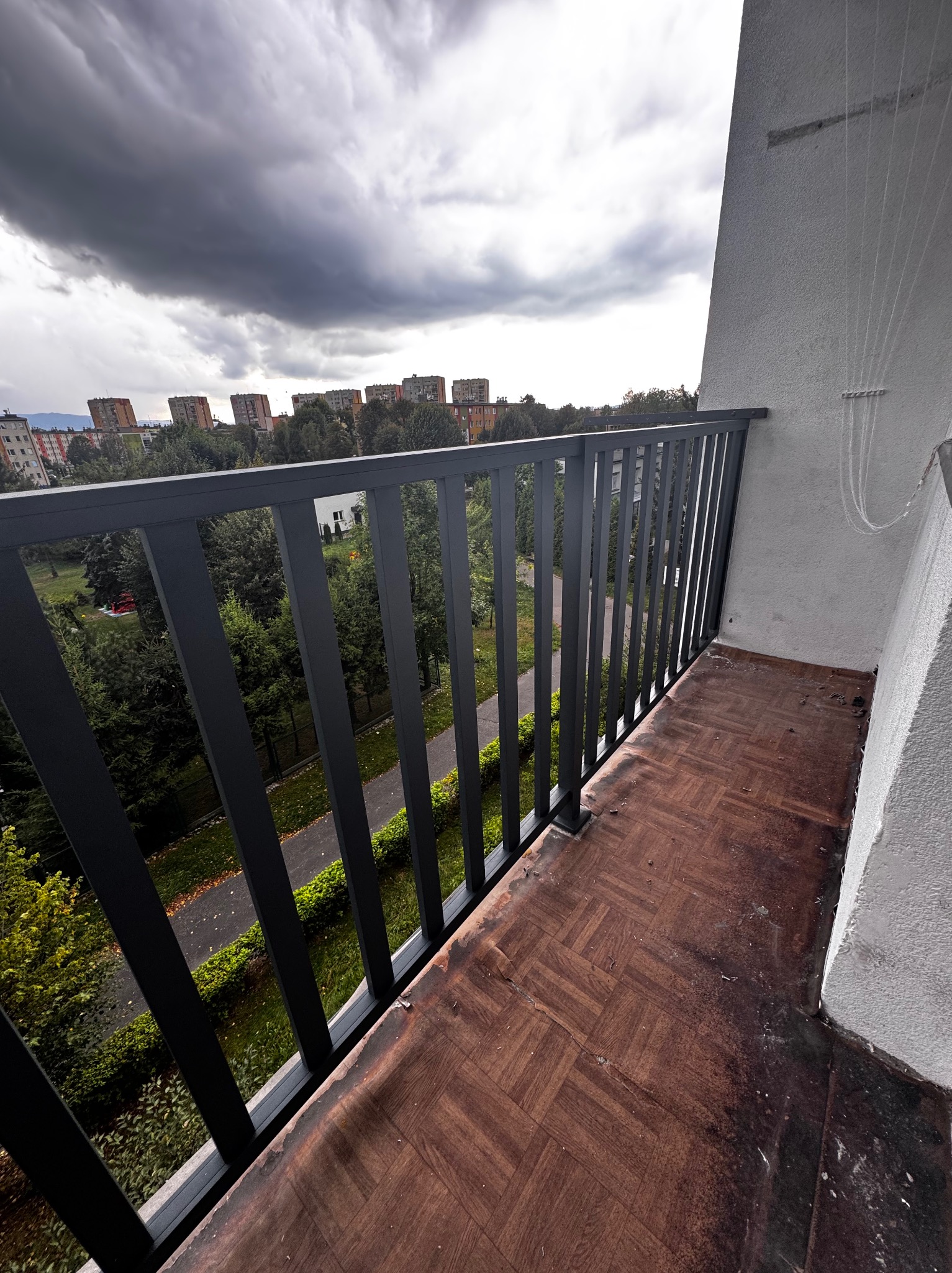 Balkon z metalową balustradą w kolorze grafitowym, widok na zieleń miejską i zachmurzone niebo. Podłoga wyłożona brązową płytką imitującą parkiet.