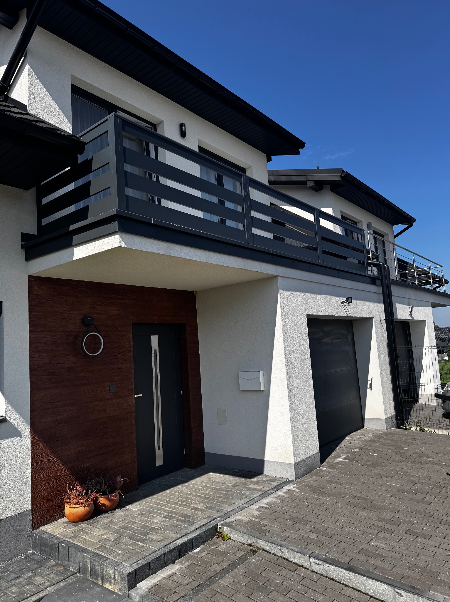 Nowoczesny dom z balkonami z czarnymi, poziomymi balustradami. Widoczne grafitowe drzwi wejściowe i bramy garażowe. Elewacja biała, tło stanowi błękitne niebo.