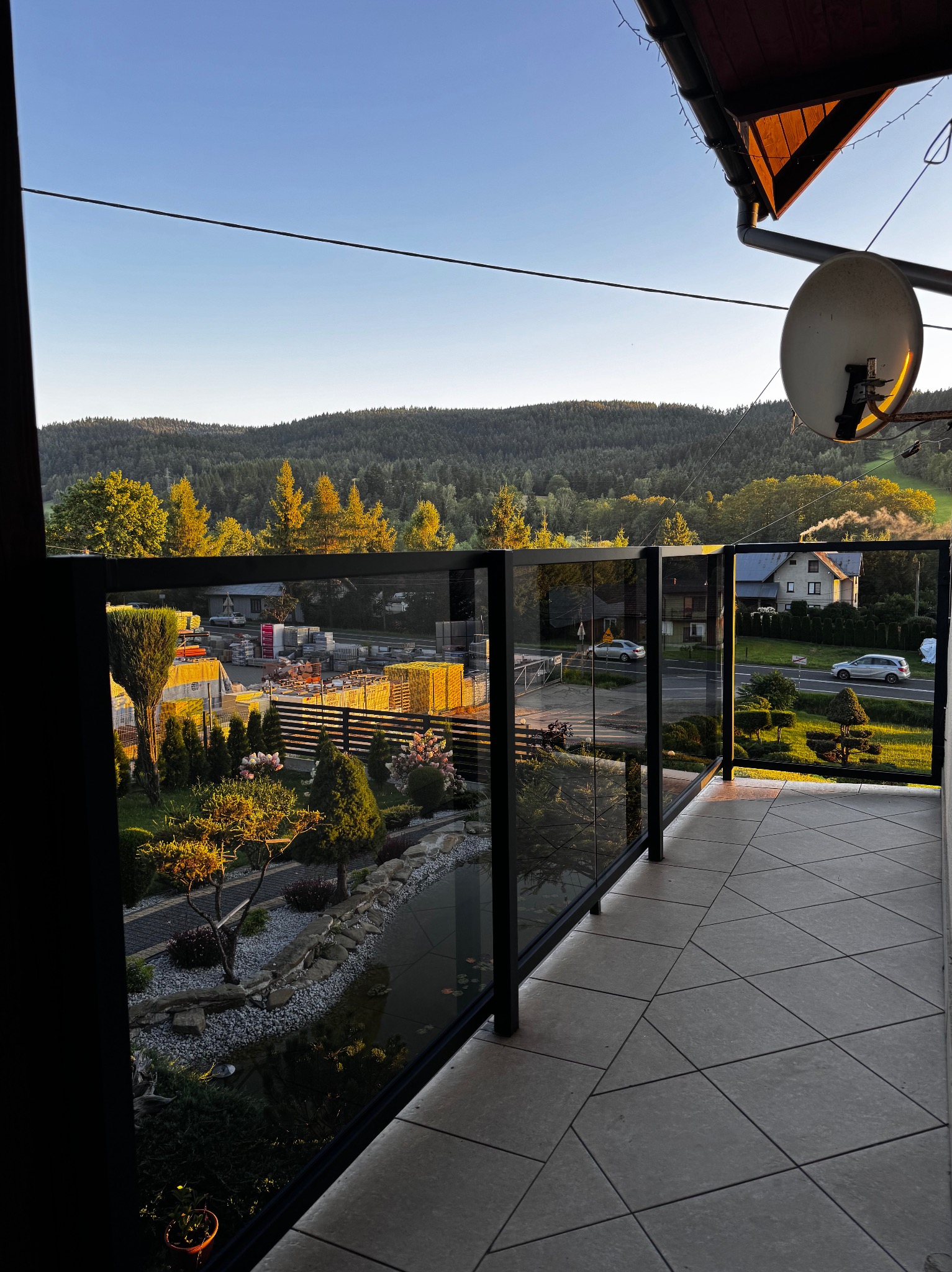 Szklana balustrada balkonowa z czarnymi ramami, widok na malowniczy krajobraz zalesionych wzgórz w ciepłym świetle wczesnego poranka.