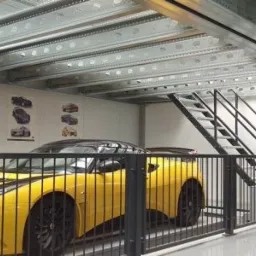 Żółty sportowy samochód wewnątrz hali z metalową konstrukcją stropu i schodami, plakaty z samochodami na ścianie.