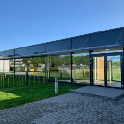 Nowoczesny budynek z przeszkloną fasadą i dwuskrzydłowymi drzwiami wejściowymi, widoczny trawnik i ogrodzenie, błękitne niebo w tle.