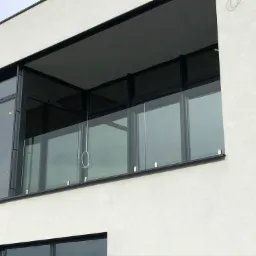 Nowoczesny balkon z przeszkloną balustradą, czarne ramy, biała elewacja budynku, widoczne mocowania szkła.