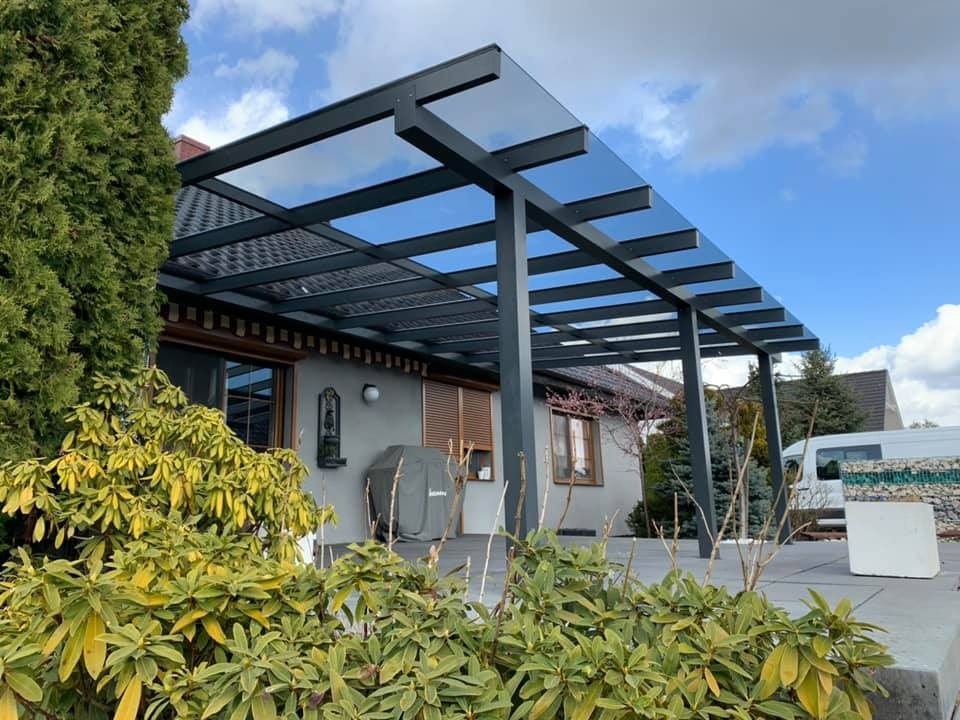 Nowoczesna, szara pergola z przezroczystym zadaszeniem, przylegająca do domu z widoczną elewacją, roletami zewnętrznymi i elementami dekoracyjnymi. Na pierwszym planie bujna roślinność, w tle...
