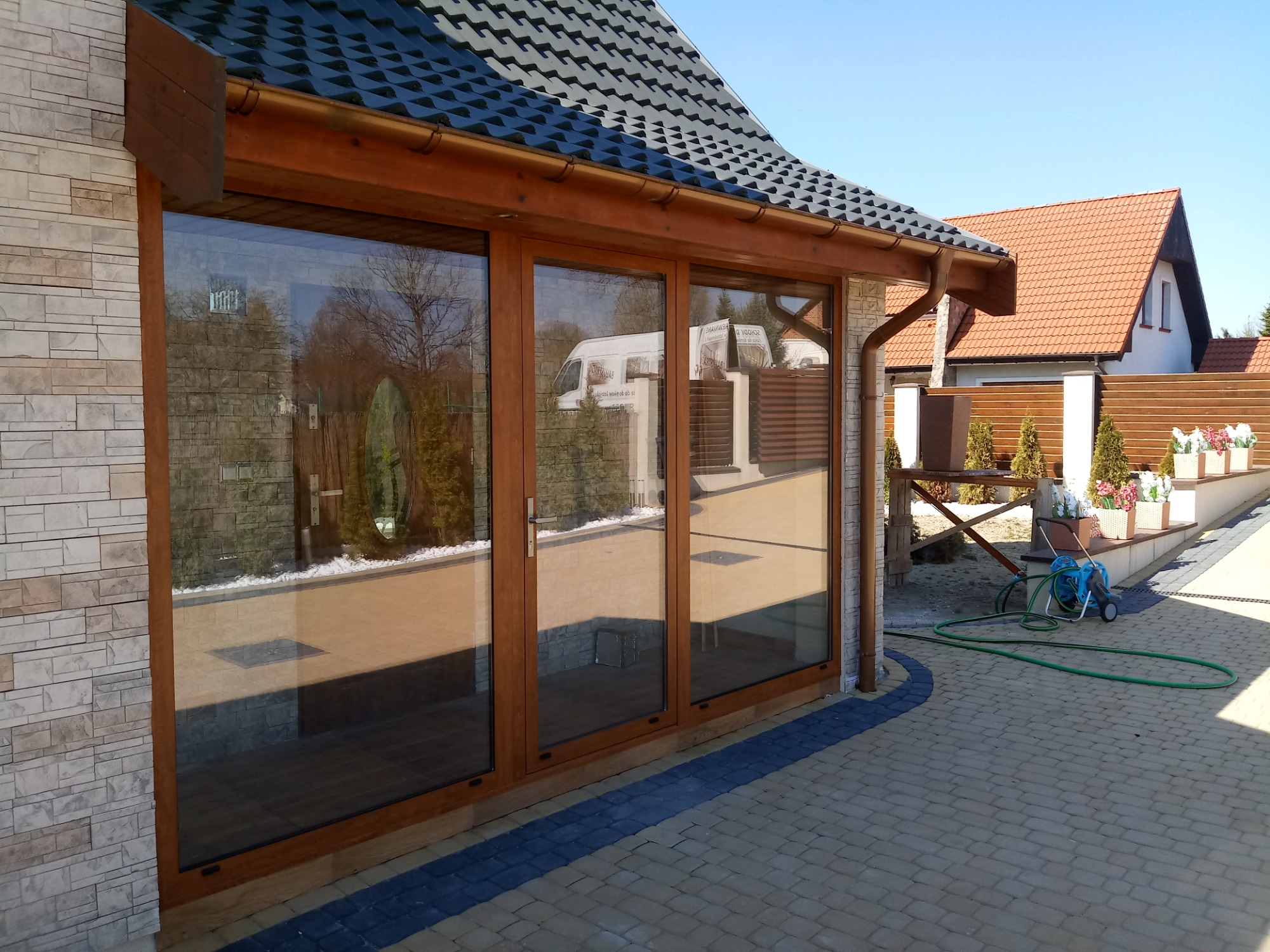 Duże, trzyskrzydłowe okno z brązowymi ramami, odbijające w szybach krajobraz ogrodu i zaparkowany biały samochód dostawczy, zamontowane w elewacji budynku wykończonej kamieniem dekoracyjnym.