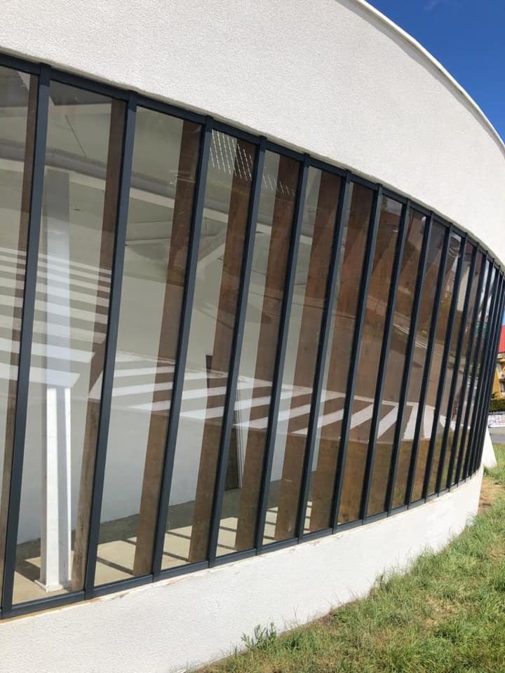 Zakrzywiona fasada budynku z szeregiem pionowych, aluminiowych ram okiennych i brązowymi panelami wewnętrznymi, widoczna z zewnątrz na tle trawnika i błękitnego nieba.