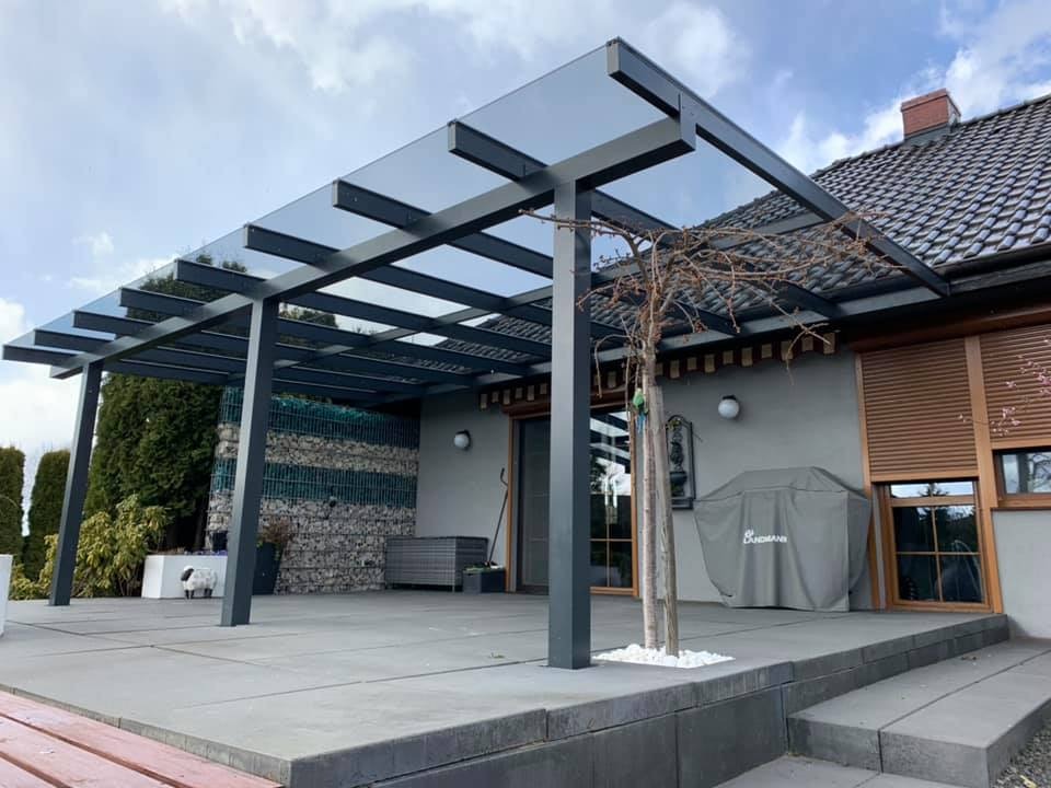 Zadaszenie tarasu z szarych profili aluminiowych i szklanym dachem, widok z zewnątrz na tle domu z szarą elewacją i brązowymi roletami, obok grill pod pokrowcem i małe drzewko.
