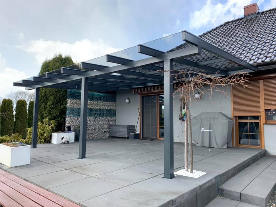 Nowoczesna, szara pergola z przeszklonym dachem na tarasie z szarych płyt, widok na dom z roletami zewnętrznymi i dekoracyjną ścianę z gabionów.