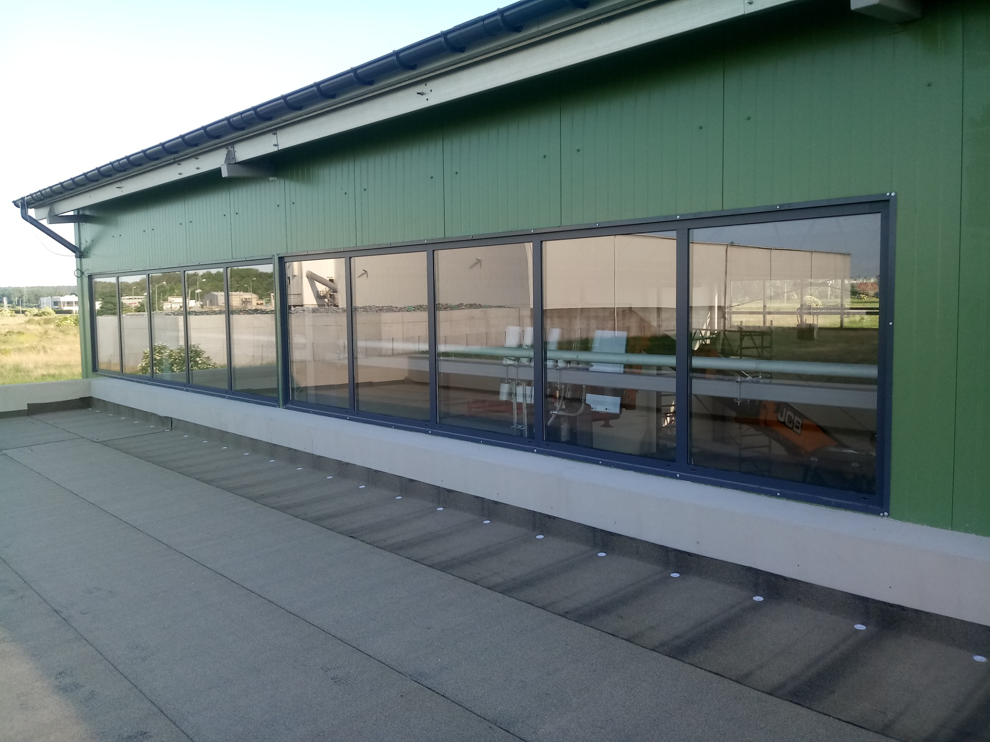 Długi szereg okien z ciemnymi ramami na tle zielonej, panelowej ściany budynku przemysłowego. W szybach odbija się wnętrze hali z widoczną maszyną budowlaną. Na pierwszym planie płaski dach pokryty...