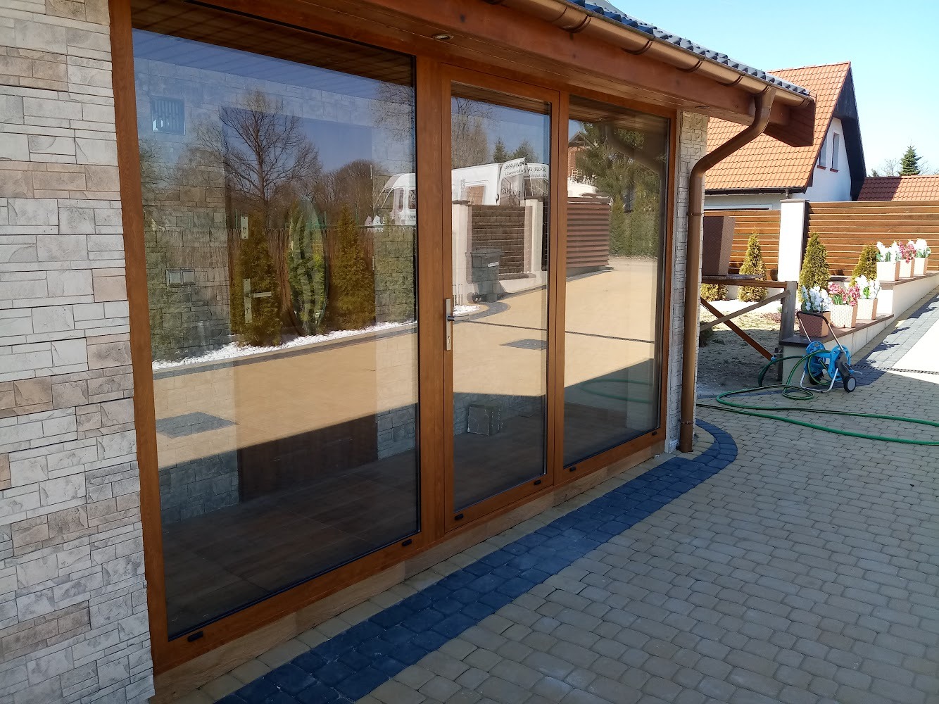 Trzyczęściowe duże okno z brązowymi ramami w elewacji domu z kamienia, widok na ogród i podjazd z kostki brukowej.