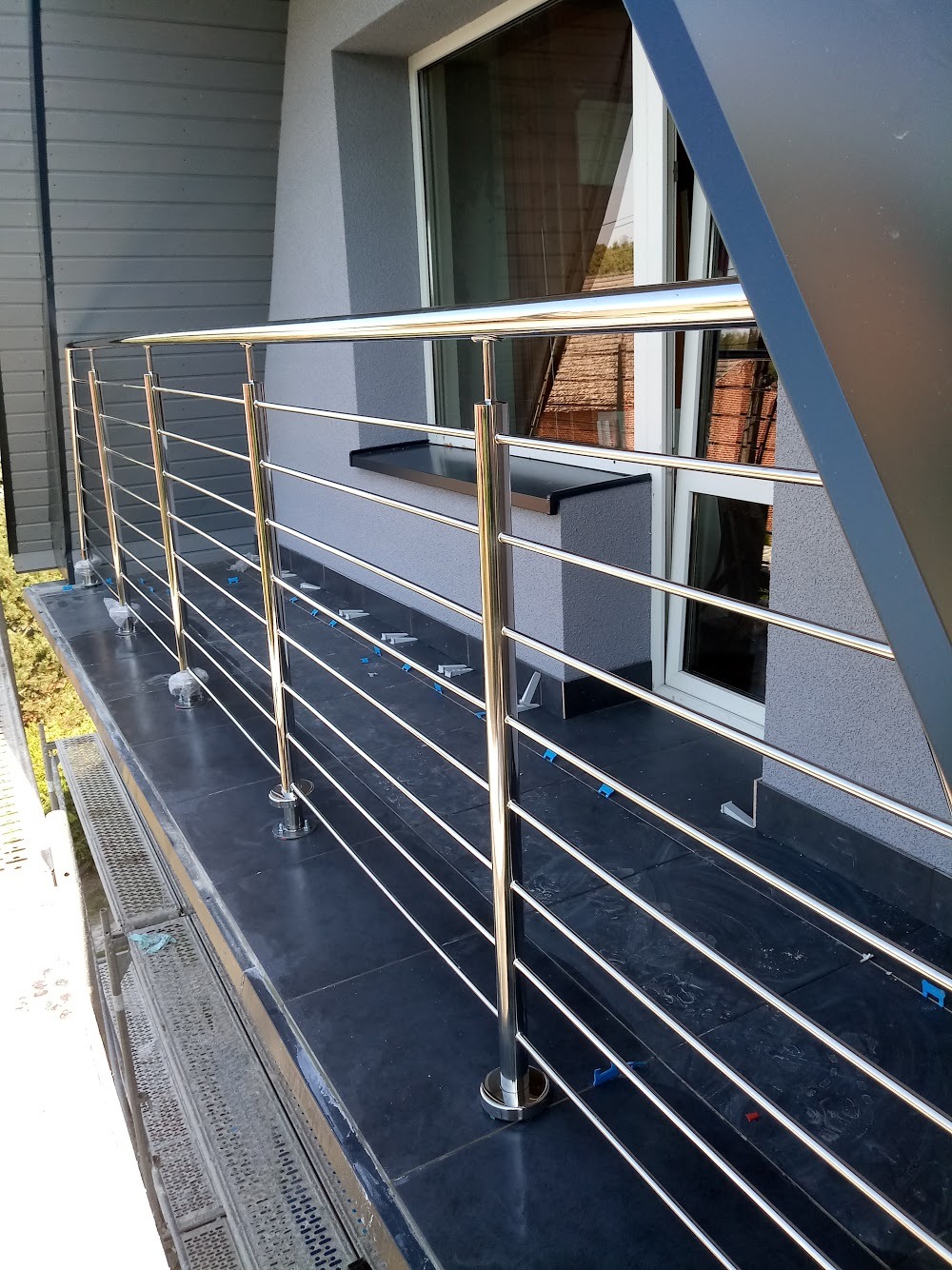 Nowoczesna, chromowana balustrada balkonowa z poziomymi prętami, zamontowana na balkonie z ciemnymi płytkami, widoczne niebieskie podkładki dystansowe pod prętami, w tle okno budynku.