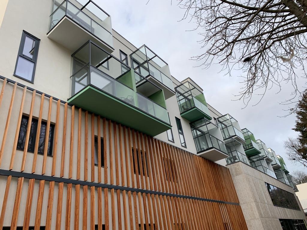 Nowoczesny budynek z balkonami z przeszklonymi balustradami i zielonymi podłogami, osłonięty drewnianymi panelami pionowymi.