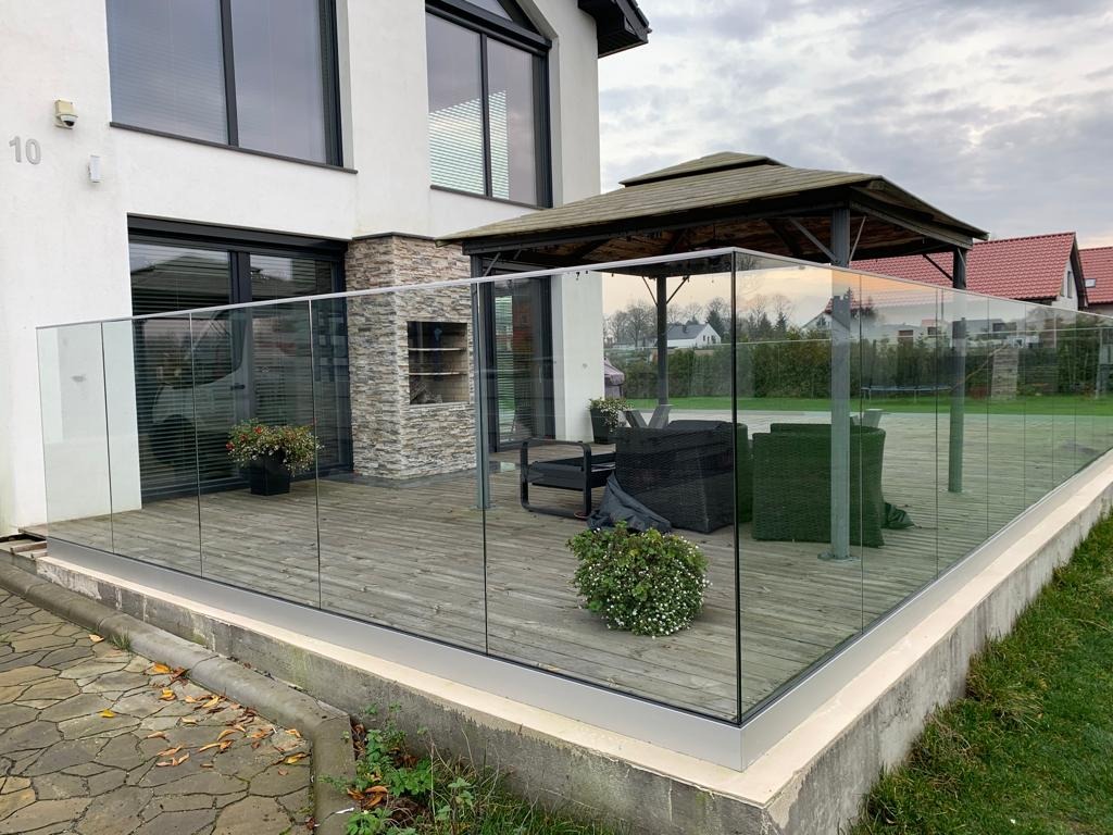 Szklana balustrada tarasowa z aluminiowymi profilami, widok na meble ogrodowe i altanę w tle.