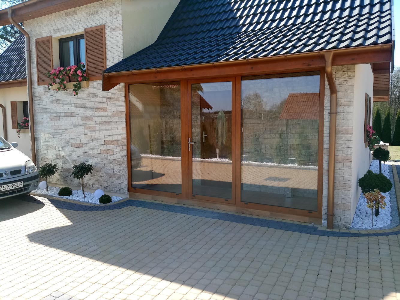 Przeszklone wejście do domu z trzema drewnianymi ramami, widok z zewnątrz na kostkę brukową i elewację z cegły, z dekoracyjnymi donicami z kwiatami w oknach.