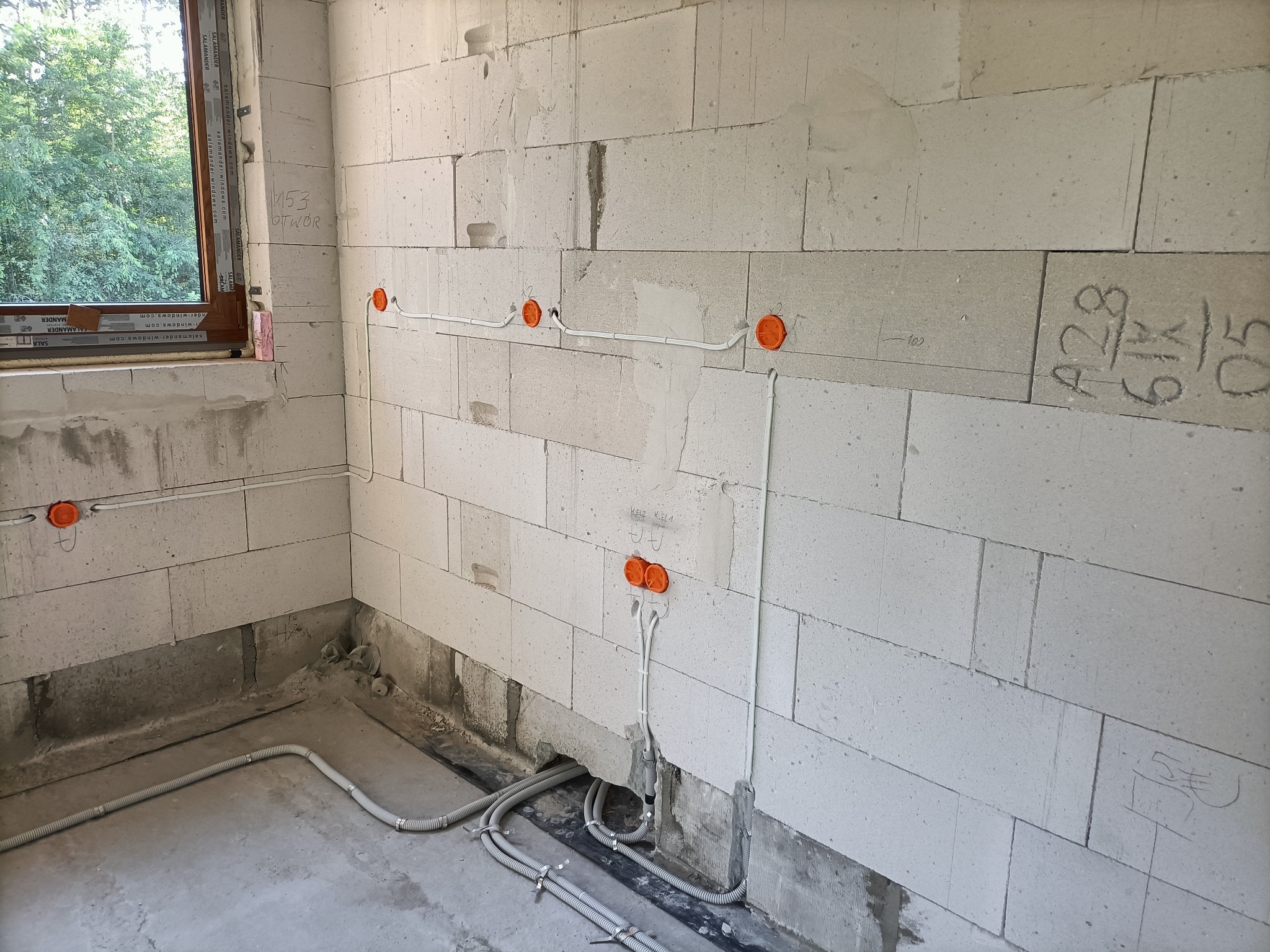 Surowy stan: Instalacja przewodów elektrycznych w ścianie z betonu komórkowego, widoczne pomarańczowe puszki instalacyjne i szare rurki karbowane przy podłodze w Józefowie.