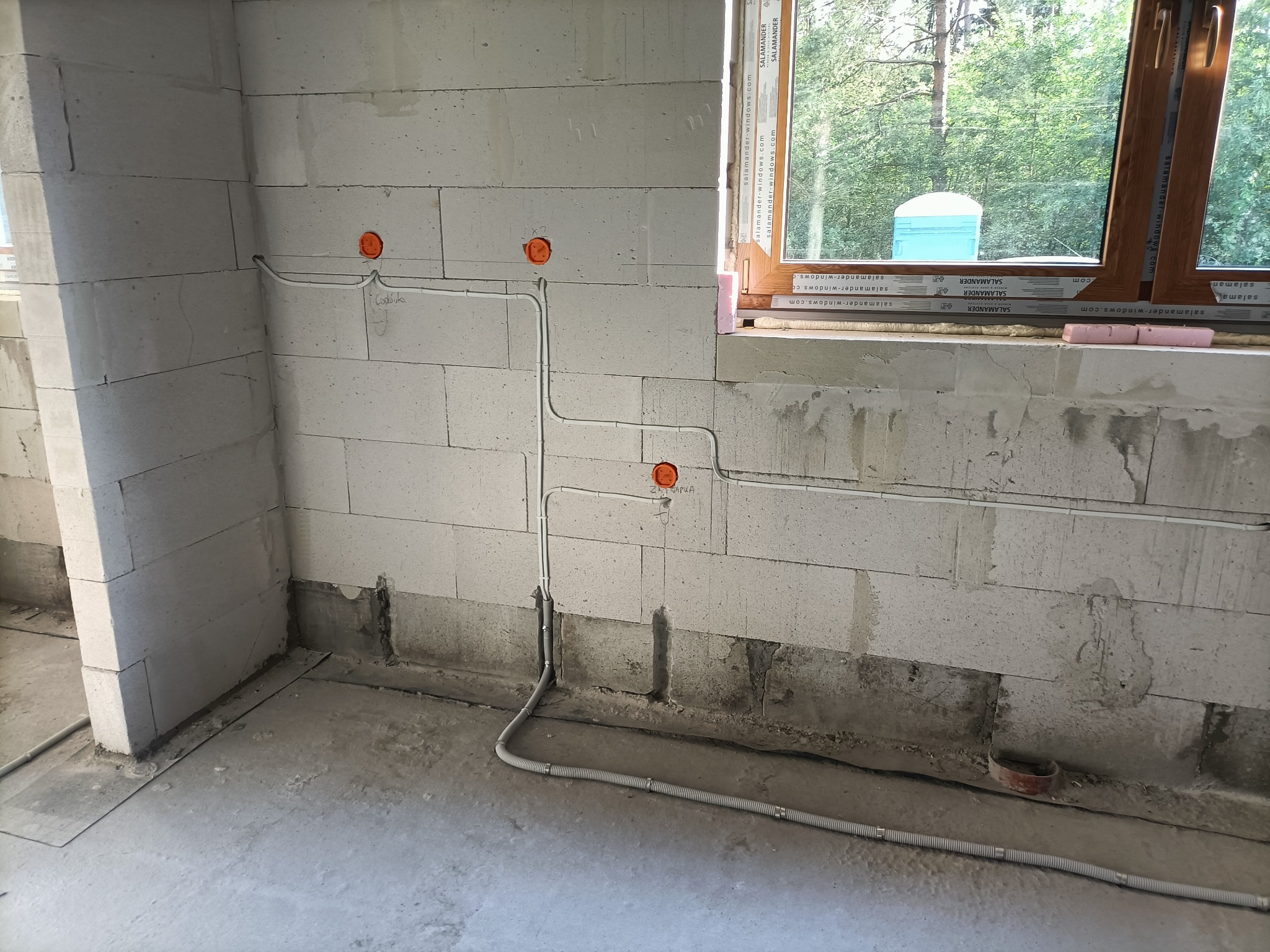 Białe ściany z pustaków, widoczne przewody elektryczne w peszlach i puszki instalacyjne z napisem 'Codouka' na ścianie, okno z widokiem na zieleń w tle.