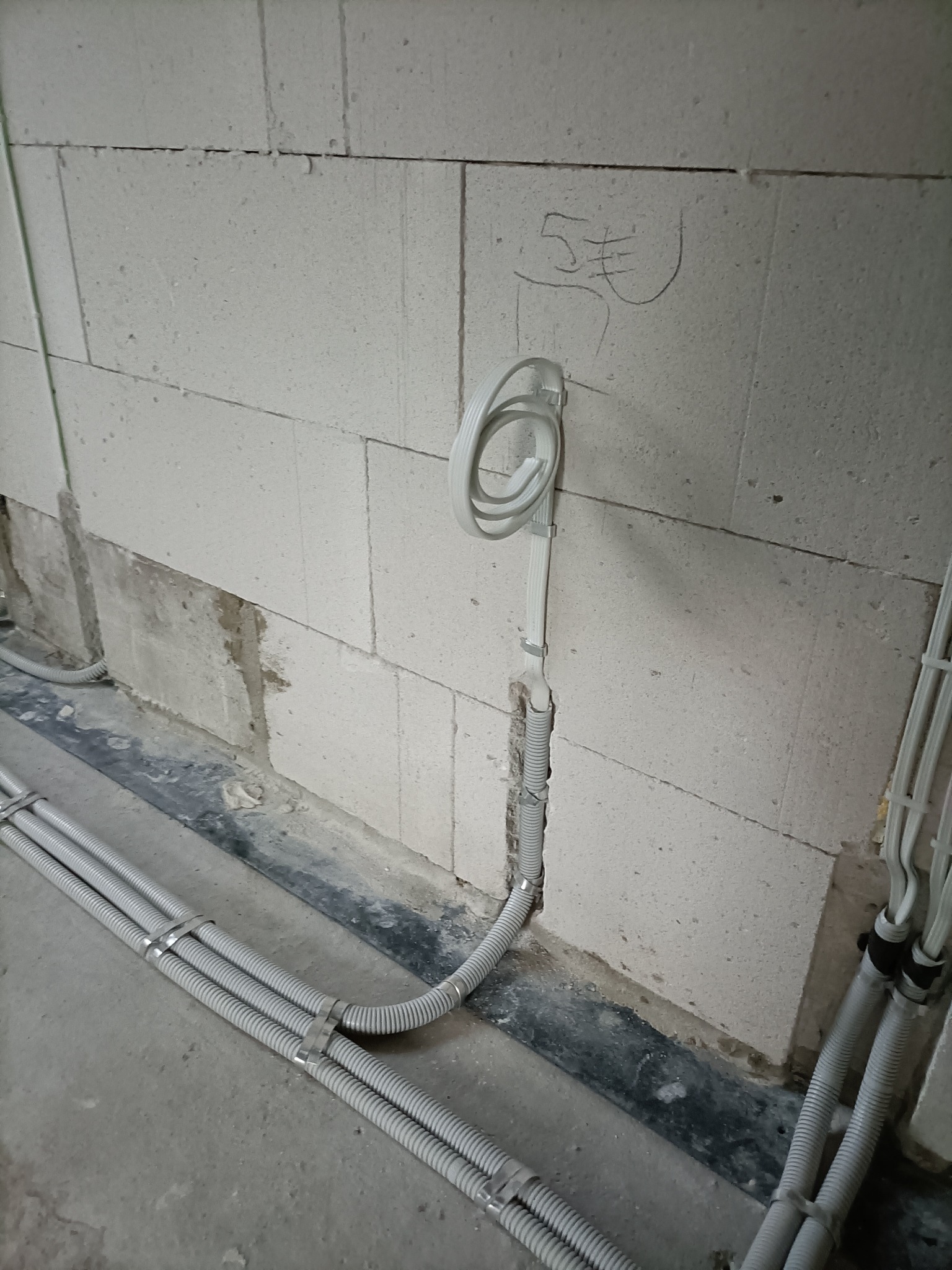 Szare kable elektryczne w peszlu prowadzone w bruździe w ścianie z betonu komórkowego, widoczne oznaczenia ołówkiem na ścianie.