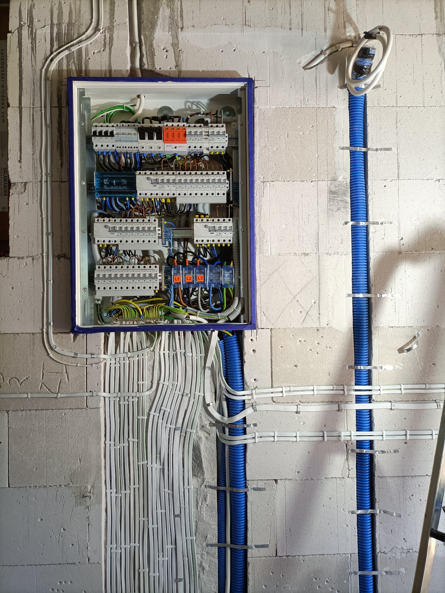Rozdzielnica elektryczna z zainstalowanymi bezpiecznikami i przewodami, umieszczona na ścianie z betonu komórkowego, z widocznymi niebieskimi rurami karbowanymi i białymi kablami mocowanymi uchwytami.