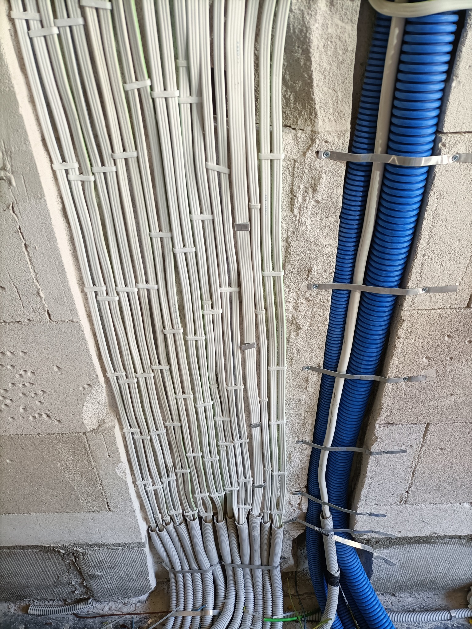 Równolegle ułożone wiązki białych kabli mocowane do ściany z betonu komórkowego za pomocą plastikowych opasek, obok niebieskie rury karbowane mocowane metalowymi uchwytami.