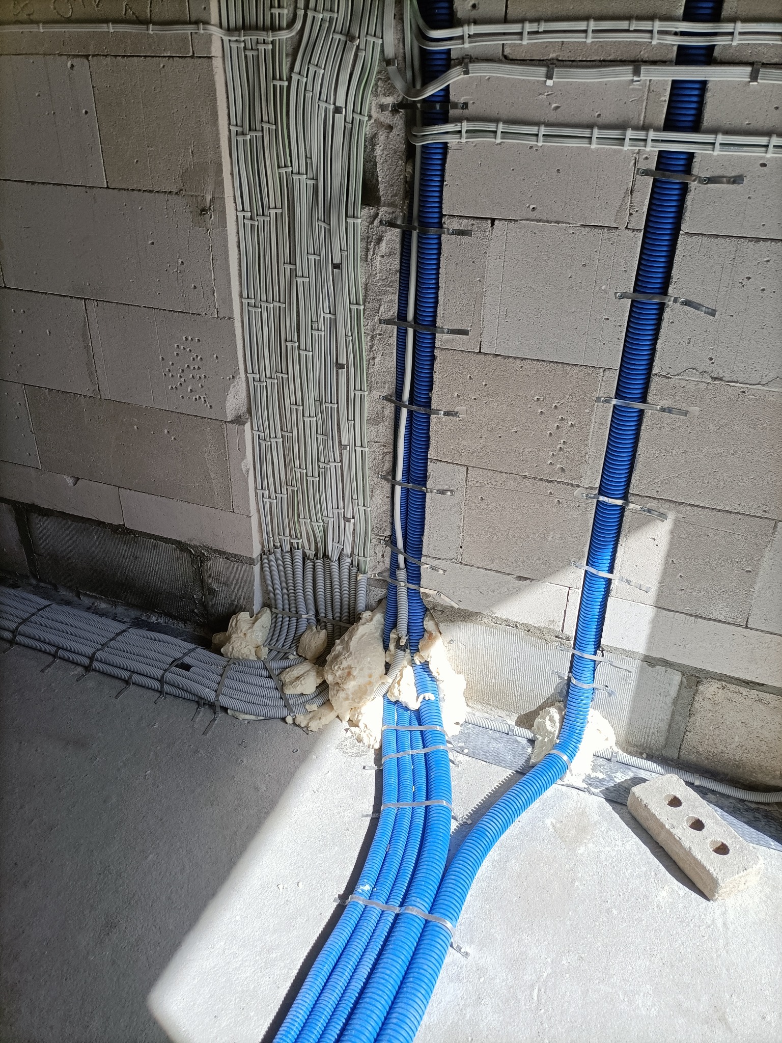 Ściana z betonu komórkowego z zamontowanymi rurami karbowanymi w kolorze niebieskim i szarym, zabezpieczonymi pianką montażową w miejscach przejść.