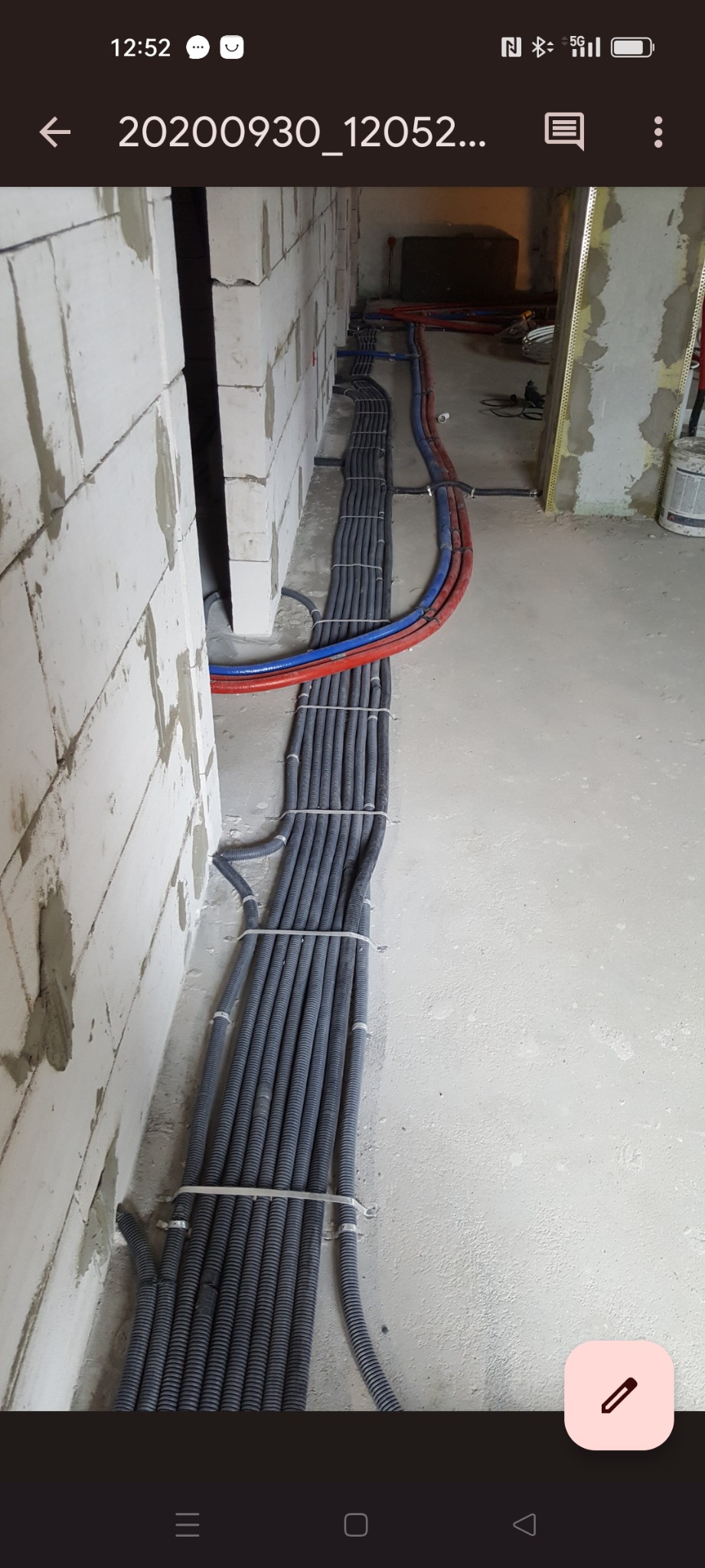 Wiązka szarych peszli z przewodami, ułożona równolegle do ściany z betonu komórkowego, zabezpieczona białymi opaskami zaciskowymi, w tle widoczne rury w kolorze czerwonym i niebieskim.