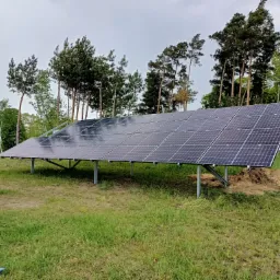 Panel słoneczny na metalowej konstrukcji, ustawiony na trawiastym terenie z drzewami w tle, widoczne ślady prac ziemnych.