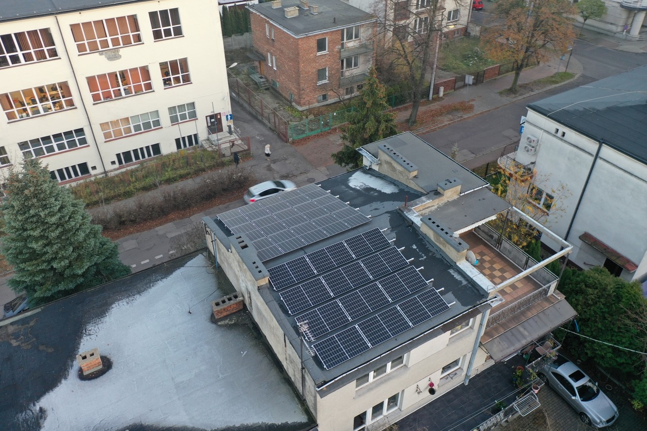 Widok z góry na budynek z panelami słonecznymi na dachu, obok zabudowa miejska, drzewa i samochody.