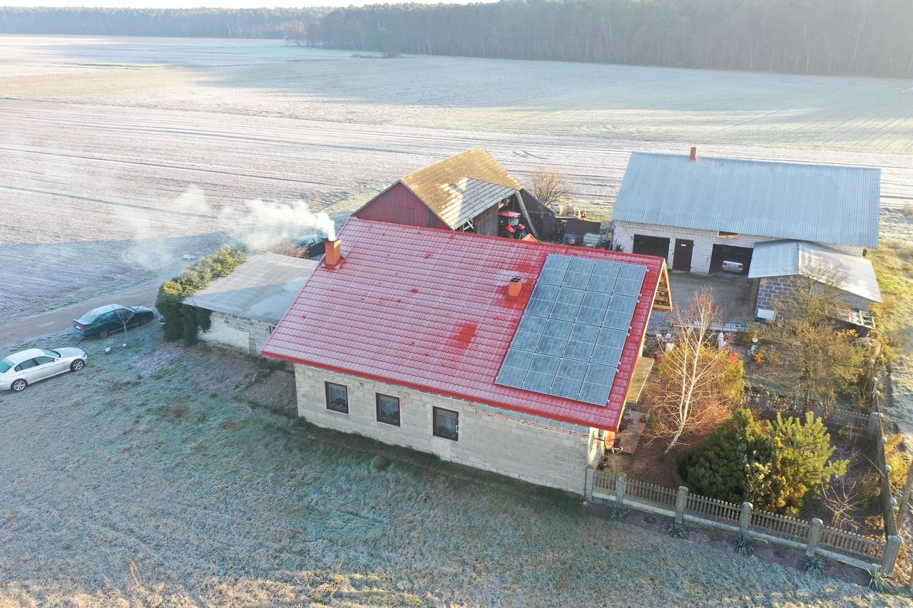 Widok z drona na dom z czerwoną dachówką i panelami słonecznymi w zimowej scenerii, z widocznymi polami pokrytymi szronem i lasem w tle.