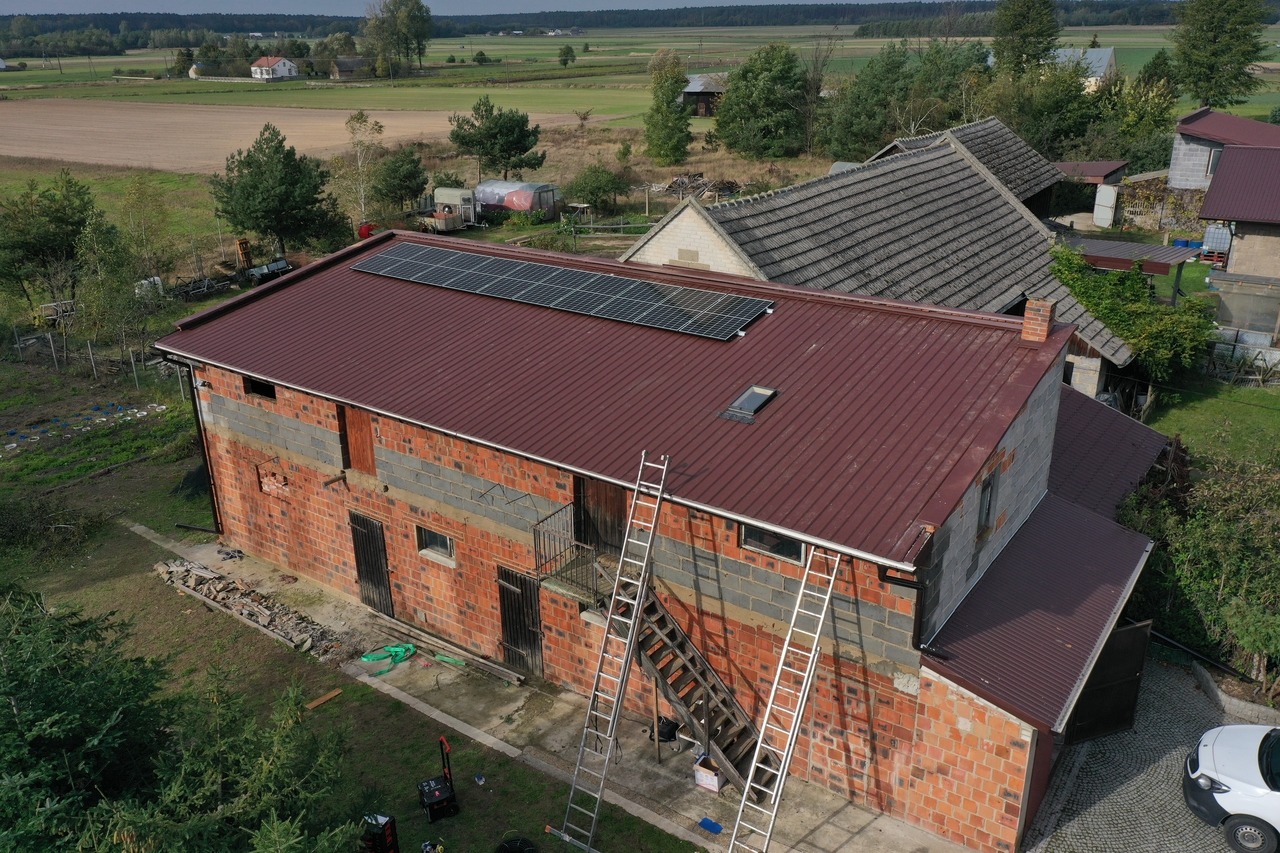 Ceglany budynek gospodarczy z nową, czerwoną blachodachówką i panelami fotowoltaicznymi na dachu, widoczne drabiny i częściowo ocieplone ściany szarego pustaka.