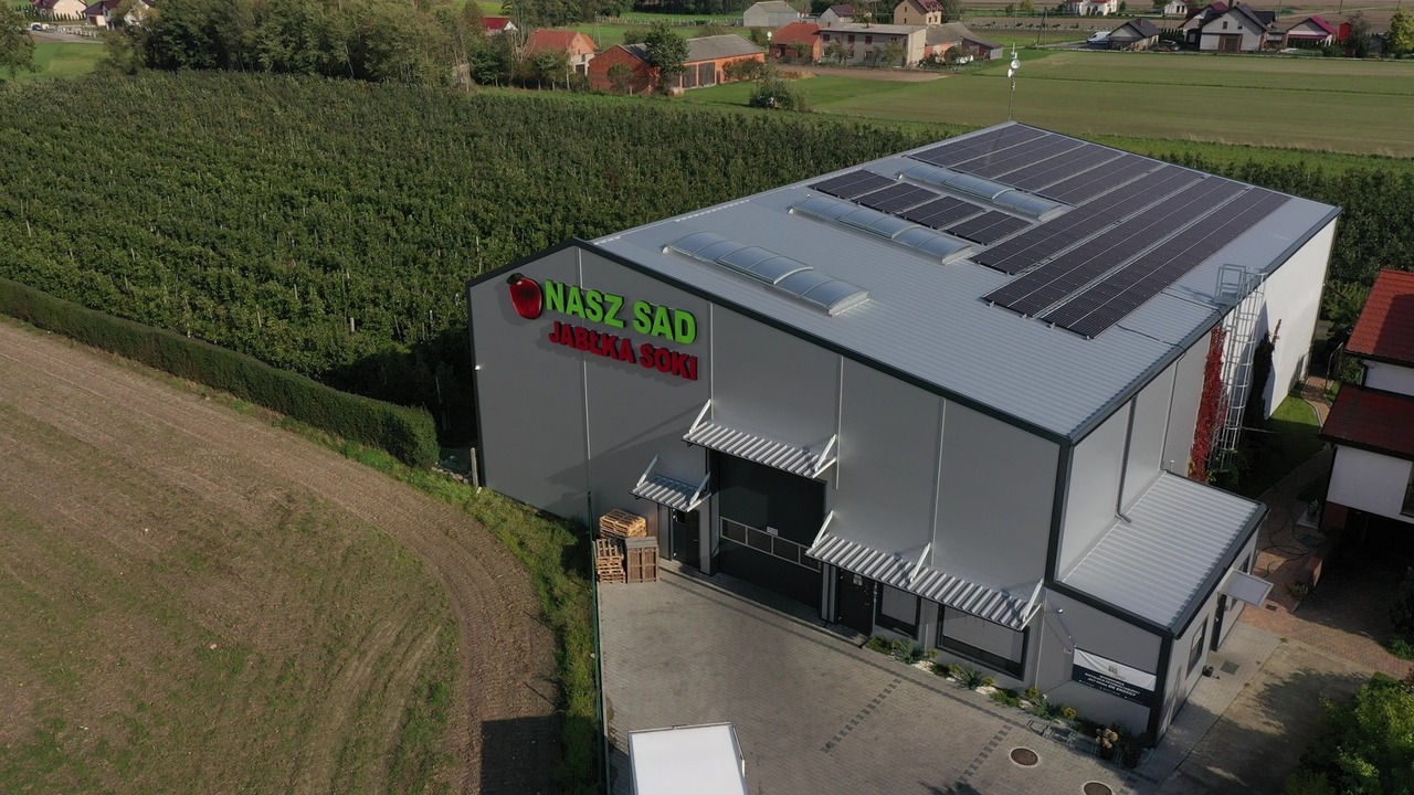 Widok z lotu ptaka na szarą halę z panelami słonecznymi na dachu, z logo 'NASZ SAD JABŁKA SOKI' i sadem jabłkowym w tle.