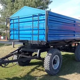 Niebieska przyczepa rolnicza z kratownicą na boku, stojąca na trawie, widoczna z boku w słoneczny dzień.