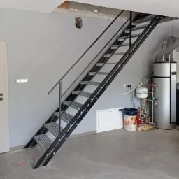 Metalowe schody ażurowe z czarną ramą i szarą kratownicą stopni, prowadzące do otworu w suficie, widoczne w surowym, szarym pomieszczeniu z instalacjami i białymi drzwiami.