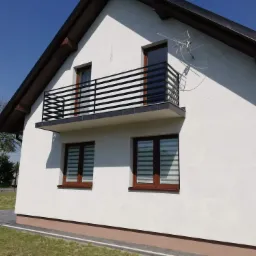 Biały dom z balkonem zabezpieczonym czarną, poziomą balustradą. Widoczna antena satelitarna na ścianie budynku.